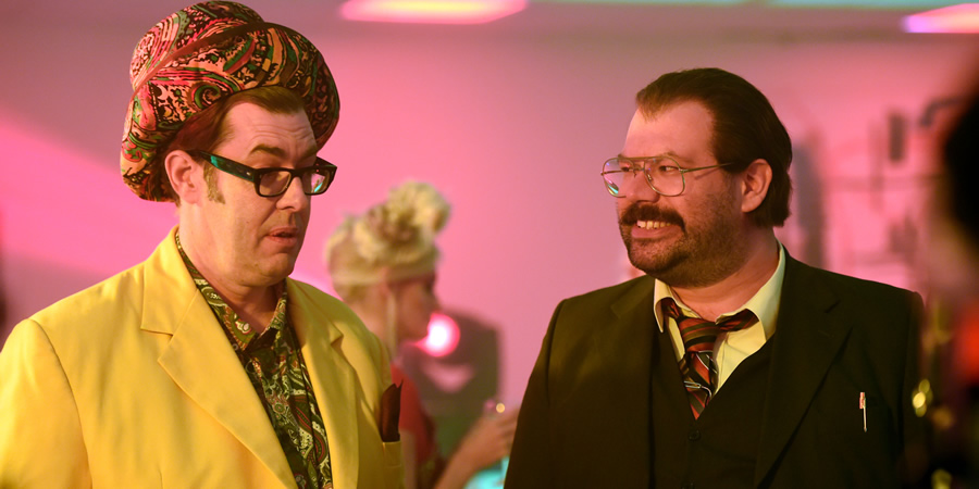 Murder In Successville. Image shows from L to R: Richard Osman, DI Sleet (Tom Davis). Copyright: Tiger Aspect Productions. Murder In Successville. Image shows from L to R: Richard Osman, DI Sleet (Tom Davis). Copyright: Tiger Aspect Productions.